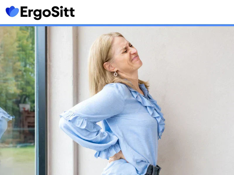 Kvinna håller sig för ländryggen med smärta, står vid ett fönster i blå blus – symboliserar ryggont vid stillasittande.