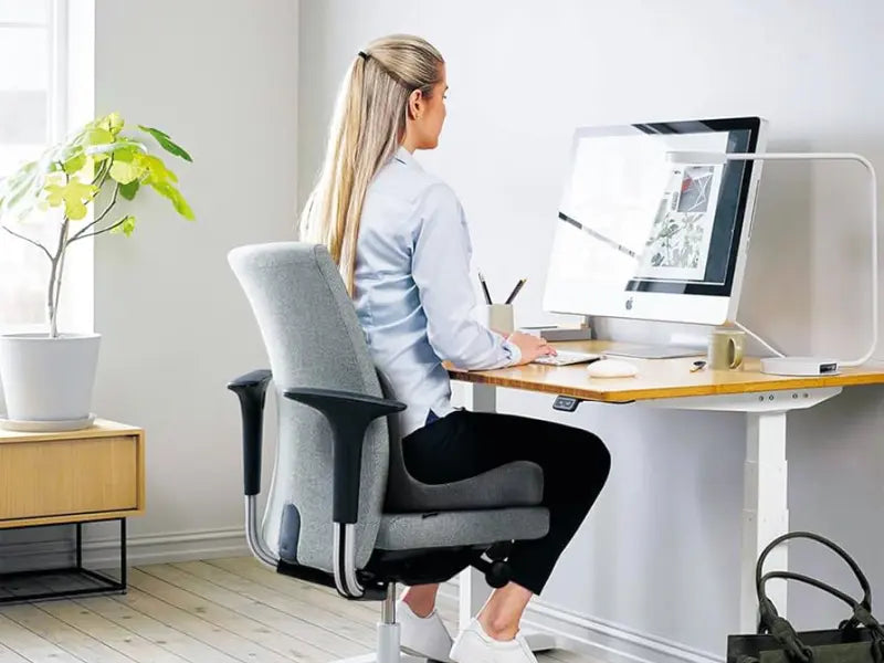 Alt-text: "Kvinna sitter ergonomiskt vid ett höj- och sänkbart skrivbord med en ergonomisk kontorsstol, framför en datorskärm i en ljus och luftig kontorsmiljö.