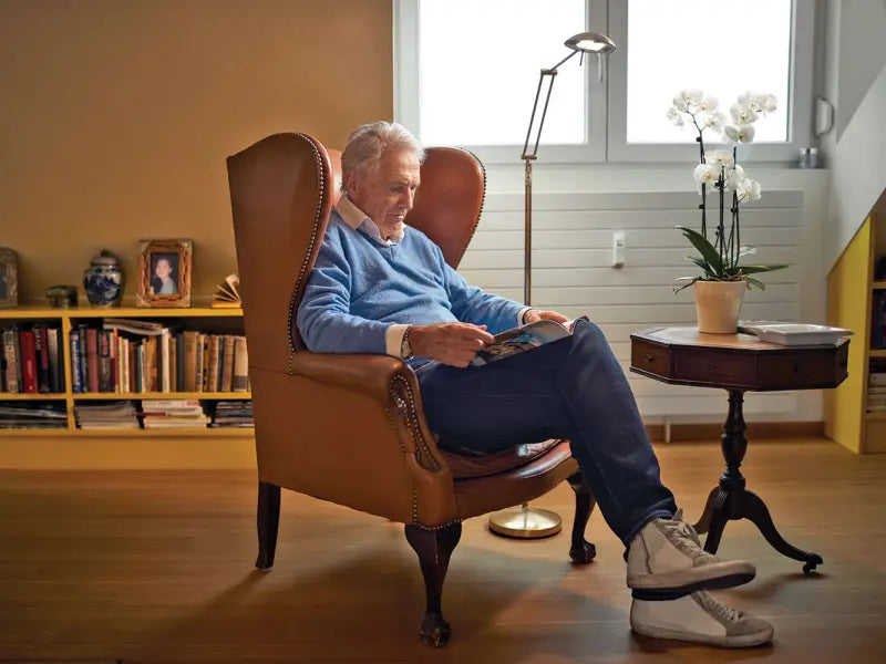 Äldre man sitter bekvämt i en fåtölj och läser en tidning i ett hemtrevligt vardagsrum med bokhylla och blomma på sidobordet