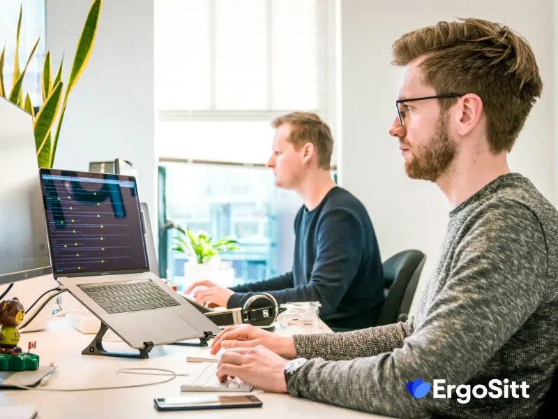 Två kontorsarbetare sitter med god hållning vid skrivbord med laptopstativ, ergonomisk arbetsmiljö och tryckavlastande sittkudde – en lösning för att förebygga ryggont vid stillasittande enligt experten Emma Nilsson på ErgoSitt.