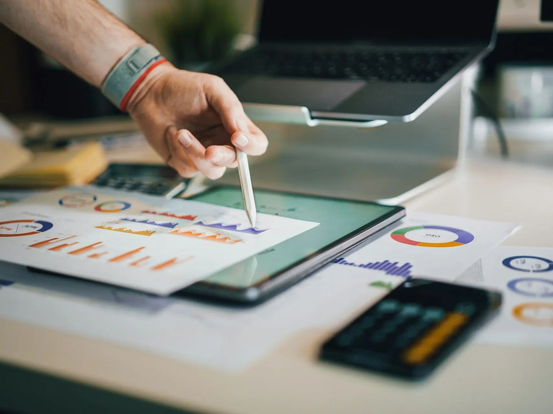 Närbild på en hand som pekar på en surfplatta med statistik och grafer, omgiven av kontorsmaterial. Bilden representerar analys av data eller affärsplanering med hjälp av digital teknik.