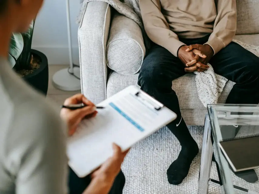 Två personer under ett samtal i en rådgivande eller terapeutisk miljö. En person sitter med händerna knäppta i knät, medan den andra personen håller ett block och antecknar.