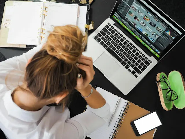 Överblicksbild av en person vid ett skrivbord, lutad över en laptop med händerna i håret, vilket indikerar stress eller frustration. På skrivbordet finns anteckningsböcker, en grön kaffemugg och andra kontorsmaterial.