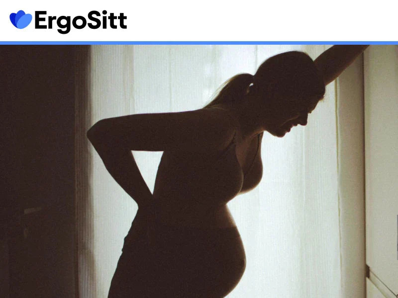 Gravid kvinna med ryggont står vid fönster och håller svanken – symboliserar behovet av ergonomiskt stöd vid sittande under graviditet
