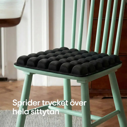 ErgoCell sittkudde i svart placerad på grön pinnstol i hemmiljö — sprider trycket över hela sittytan för bekvämare sittande