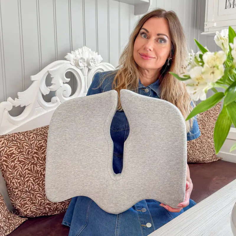 Kvinna visar upp ErgoKudden ergonomisk sittkudde i grått med avlastande urtagning för svanskota — sittande i soffa i hemmiljö