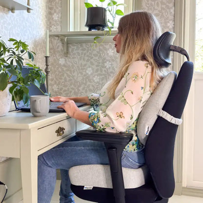 Kvinna arbetar vid skrivbord med ErgoPaket Pro — ErgoKudden på sitsen och ErgoStödet mot ländryggen på kontorsstol i hemmakontor
