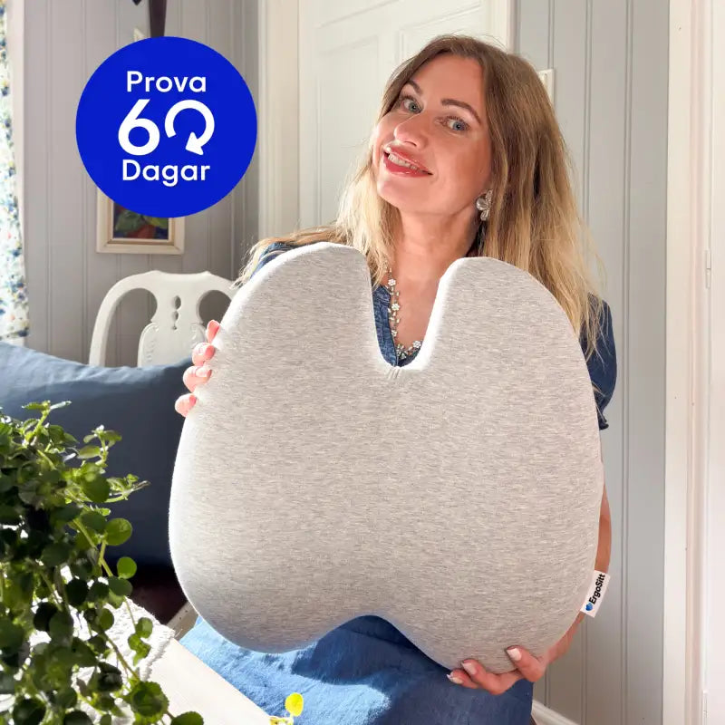 Kvinna håller upp ErgoStödet ergonomisk ländryggskudde i grått hemma i vardagsrummet — prova riskfritt i 60 dagar