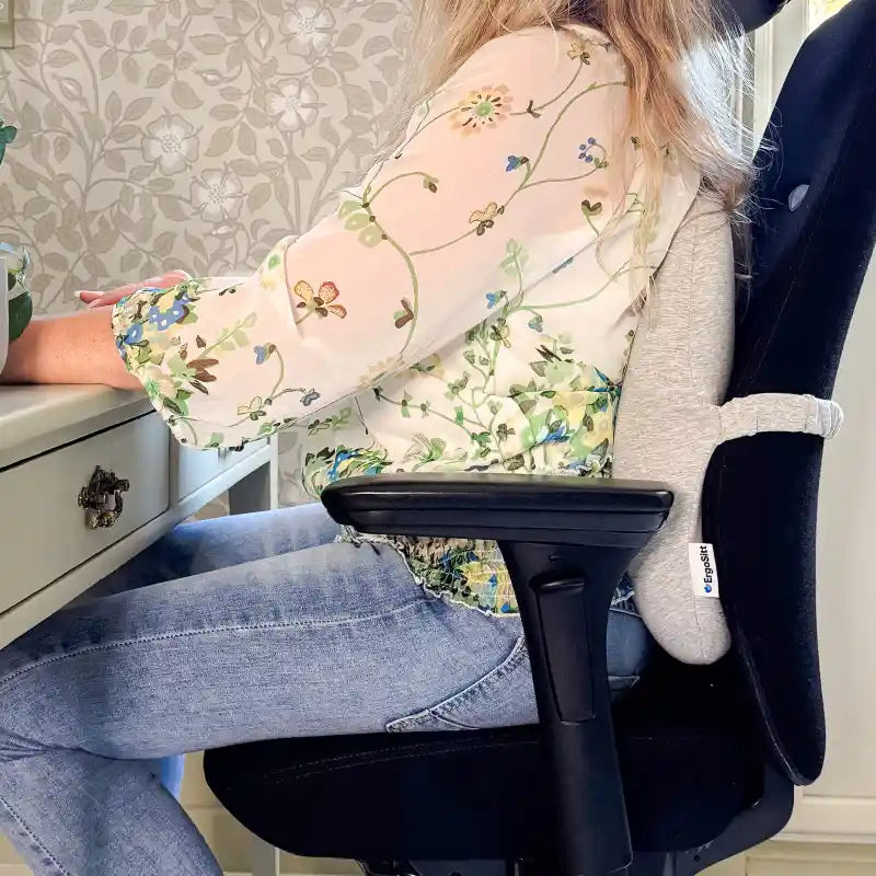 Kvinna sitter med ErgoStödet ergonomisk ländryggskudde mot nedre ryggen på svart kontorsstol vid skrivbord — stabilt ländryggsstöd under arbete